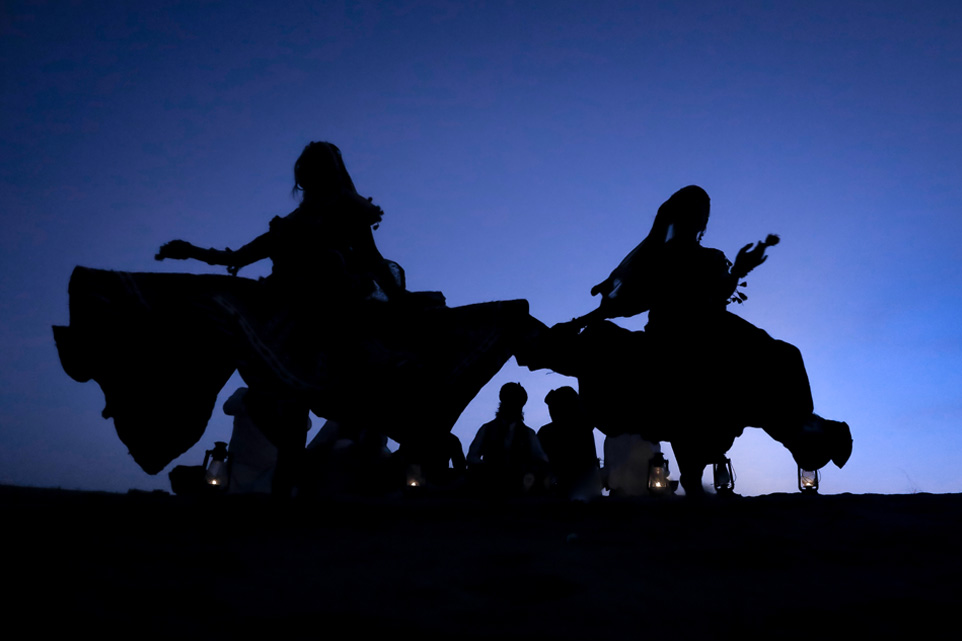 Two women dancing in flowing garments, with a dark blue sky in the background.